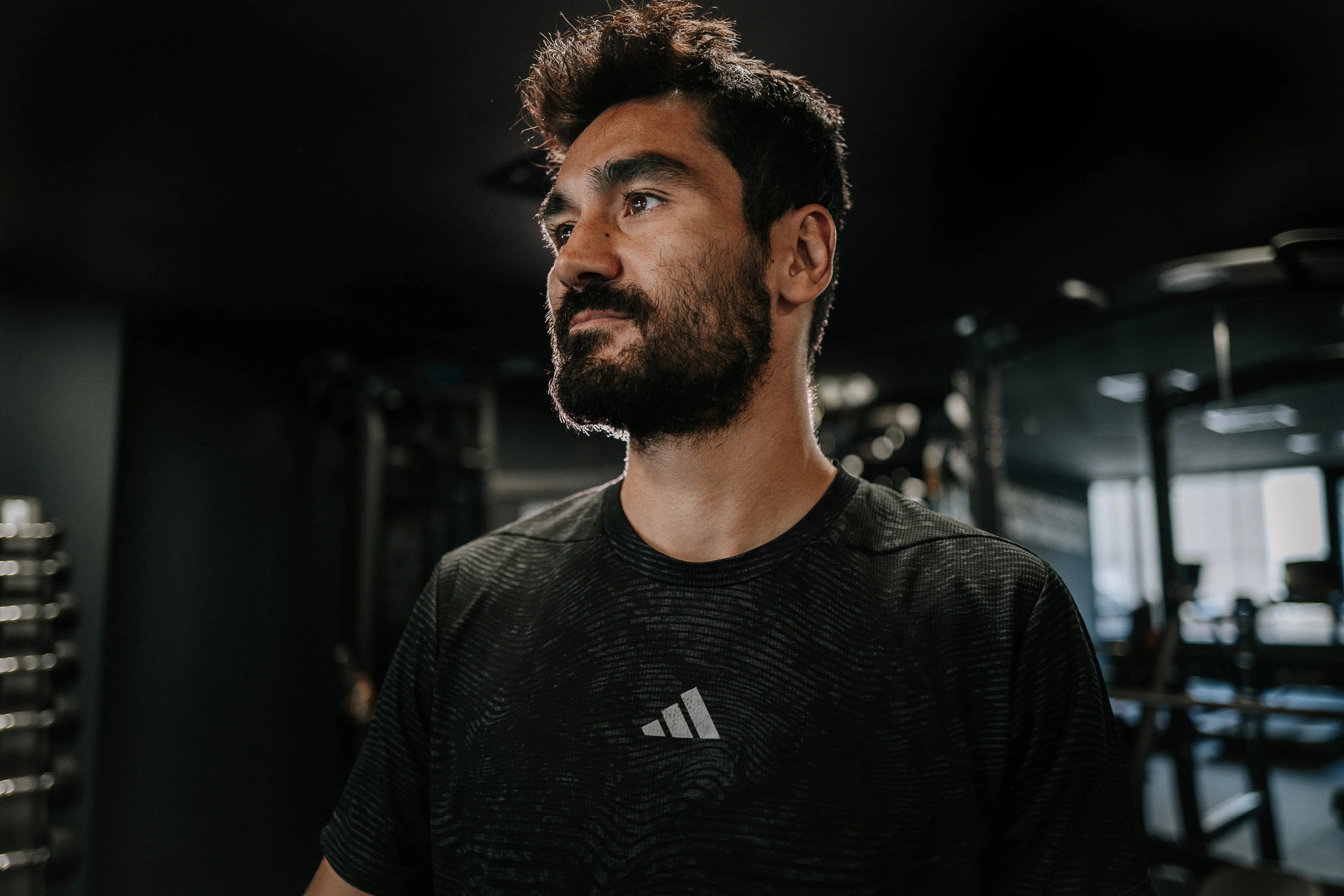 Ein Mann mit Bart und schwarzem T-Shirt steht in einem Fitnessstudio und blickt zur Seite.