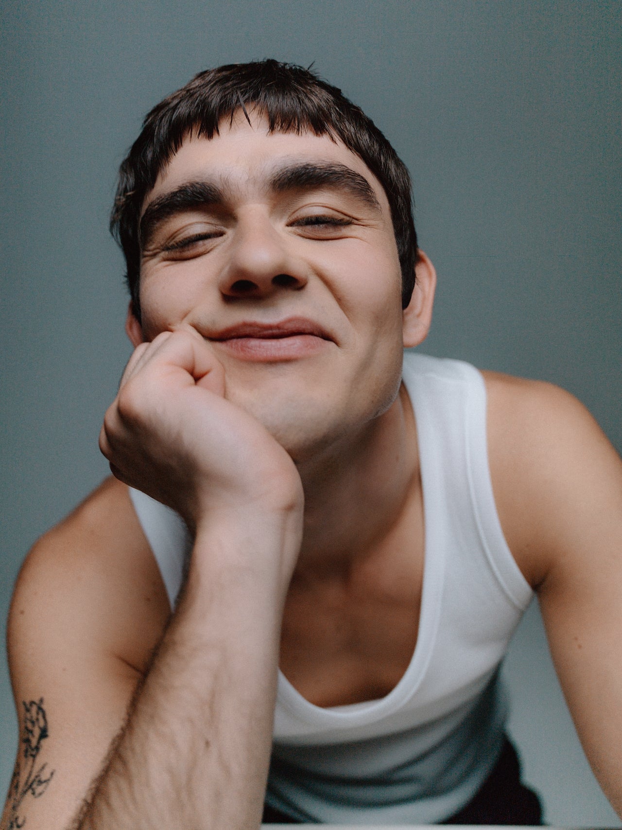 Ein lächelnder Mann im weißen Tanktop stützt sich mit der Hand am Kinn ab und schaut direkt in die Kamera.