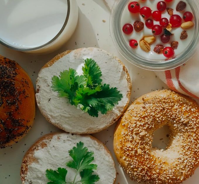 Bagels mit Frischkäse und Koriander, ein Glas Milch, und Joghurt mit roten Beeren und Nüssen auf einem hellen Tisch.
