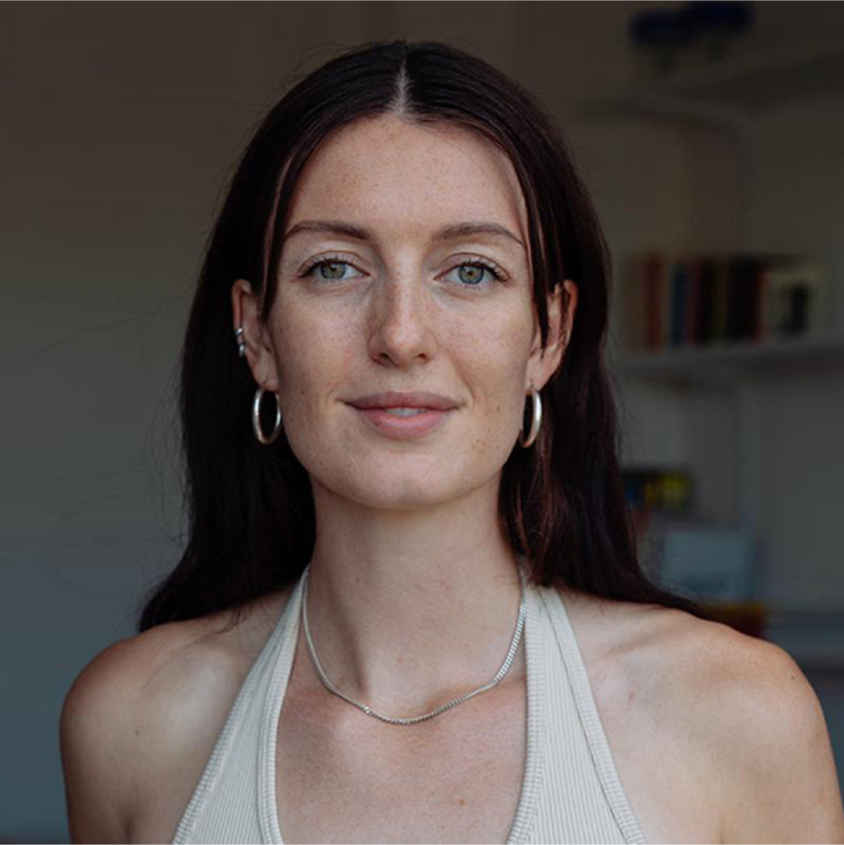 Eine Frau mit langen, dunklen Haaren und Ohrringen lächelt in die Kamera, im Hintergrund ein unscharfes Bücherregal.