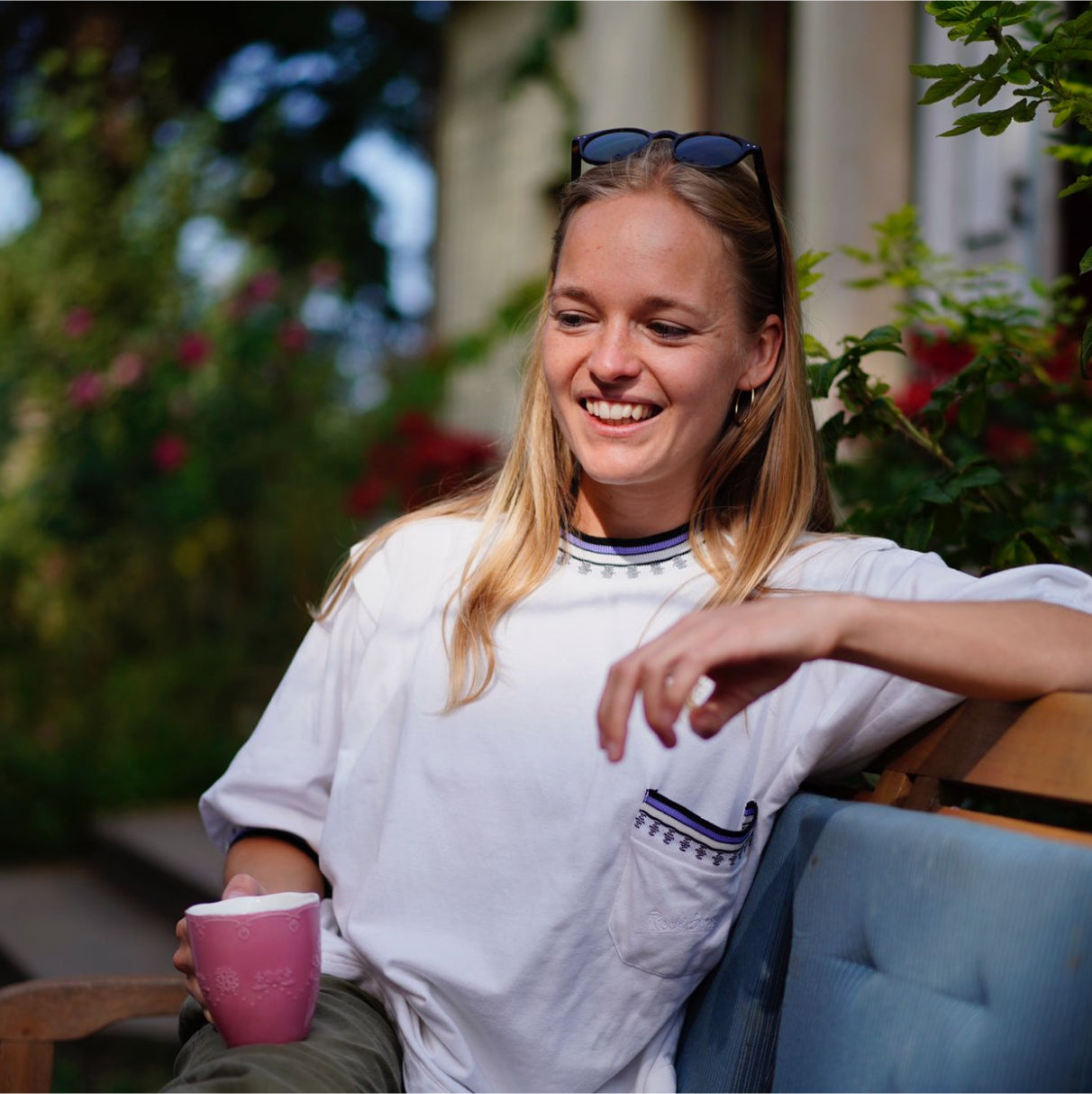 Eine Frau sitzt lächelnd auf einer Bank im Freien und hält eine rosa Tasse in der Hand.