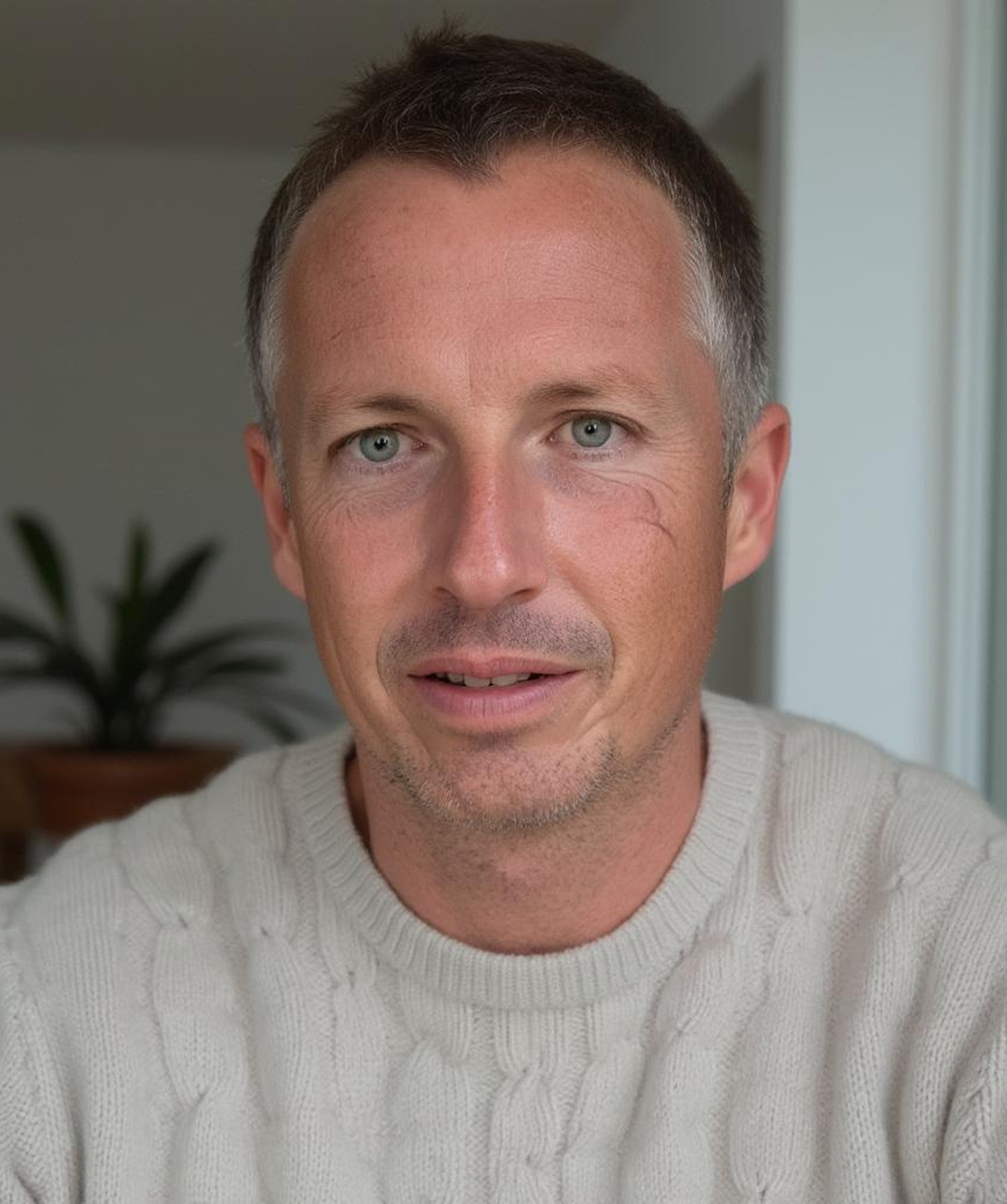 Ein Mann mit kurzen Haaren und hellem Pullover schaut in die Kamera; im Hintergrund ist eine Pflanze zu sehen.
