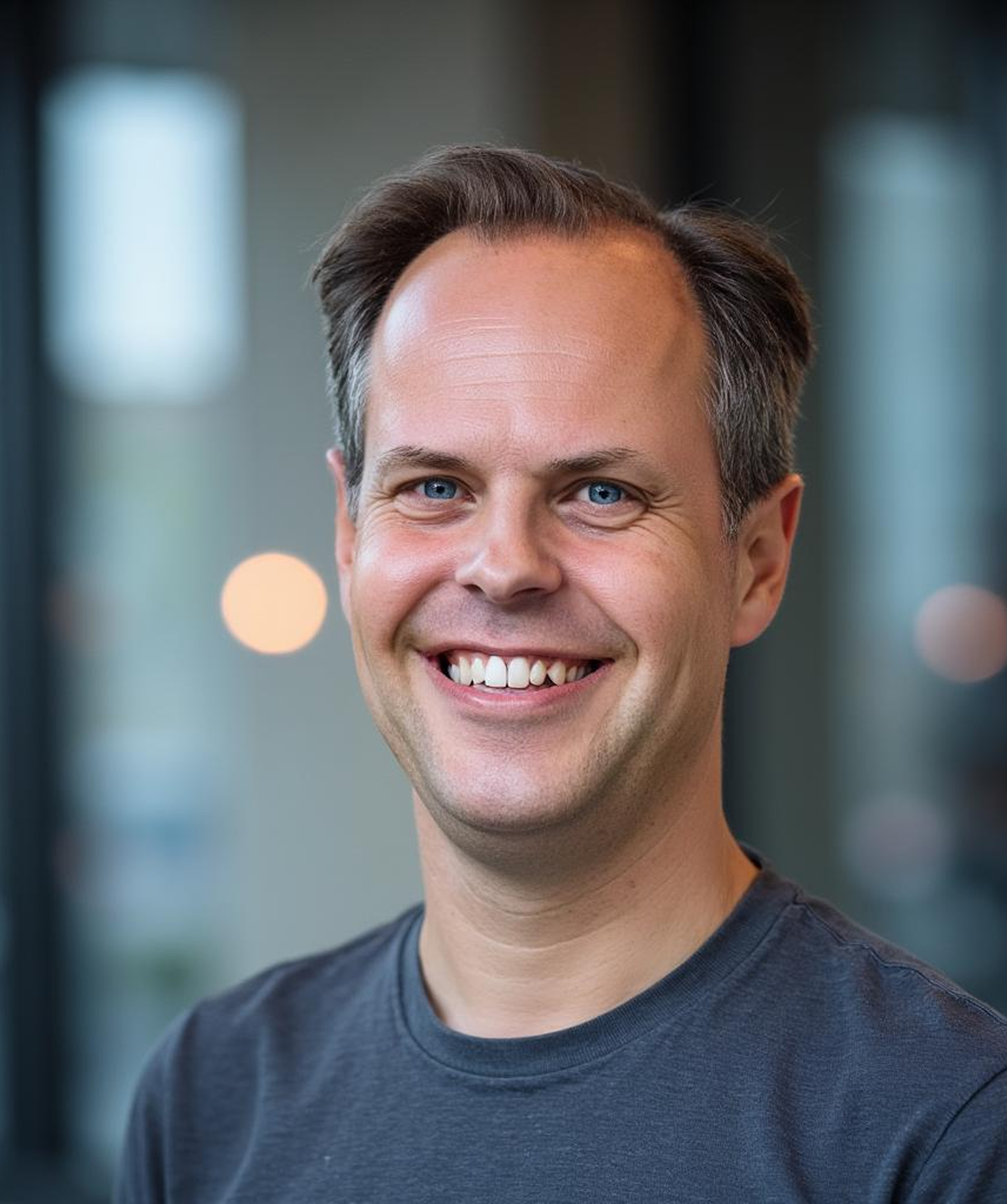 Lächelnder Mann in einem grauen T-Shirt, im Innenraum mit verschwommenem Hintergrund.