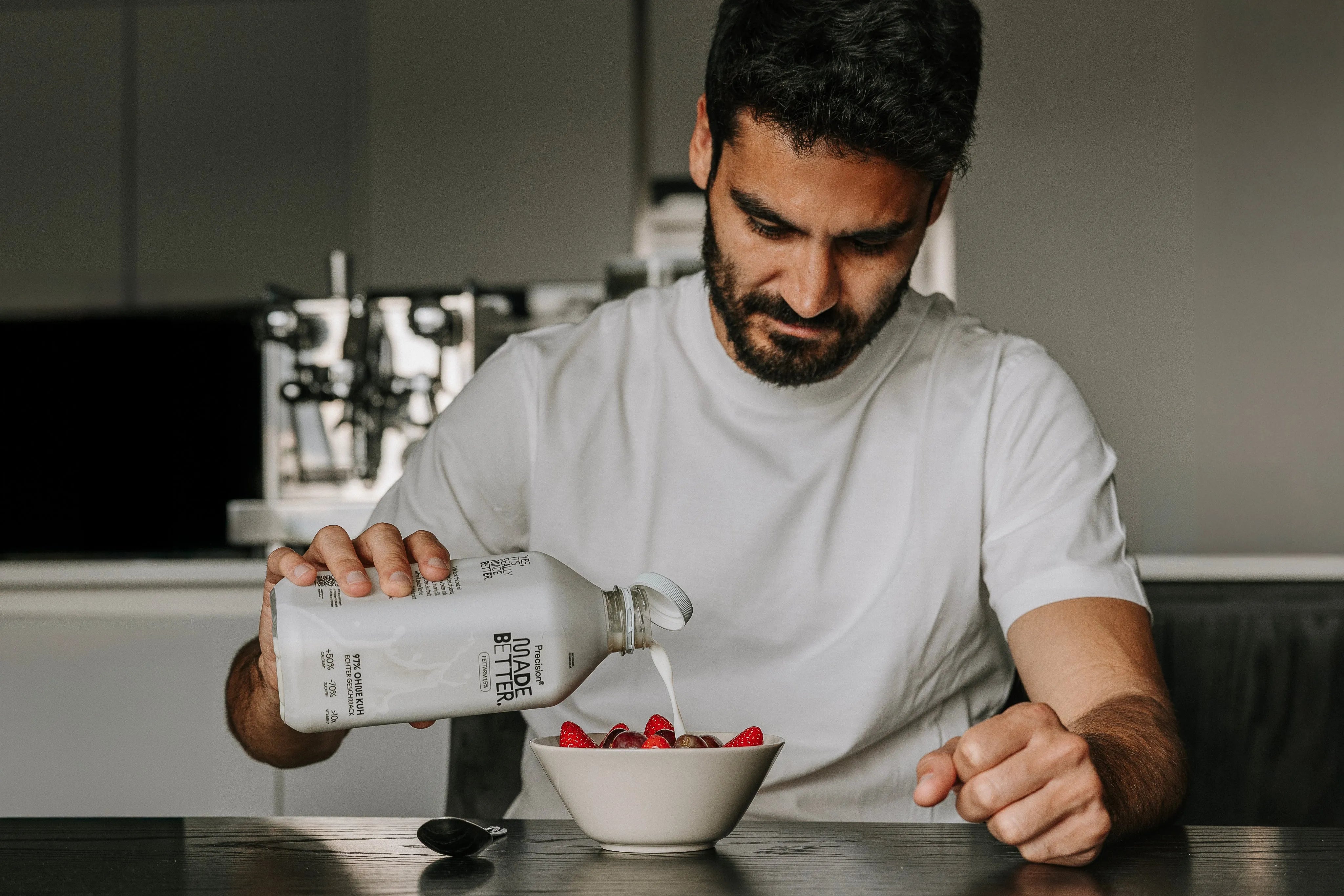 Ein Mann in einem weißen T-Shirt gießt Milch in eine Schüssel mit Beeren auf einem Tisch.